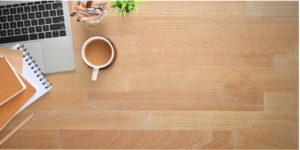 A wooden desk with a laptop, a cup of coffee, a notebook, and two brown notebooks, along with a small plant and pencils in a glass.