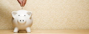A hand inserting a coin into a white piggy bank on a wooden surface, with a floral-patterned yellow background.